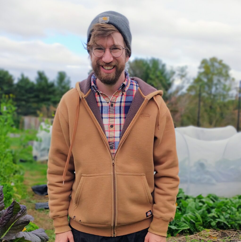 photo of Zach a white male with glasses and a beard, wearing a brown sweatshirt over a flannel shirt standing in farm. 